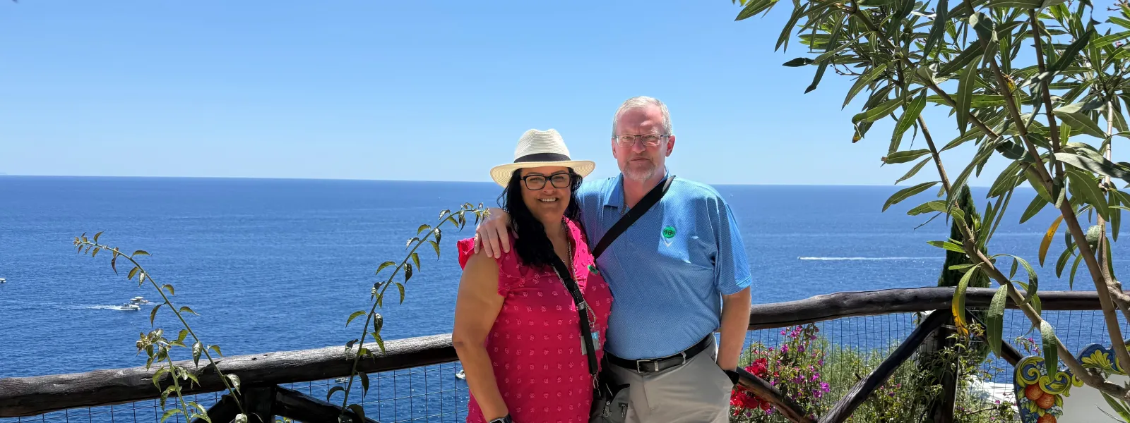 Smiling couple posing by a decorated bench overlooking the blue ocean on a sunny day with clear skies.