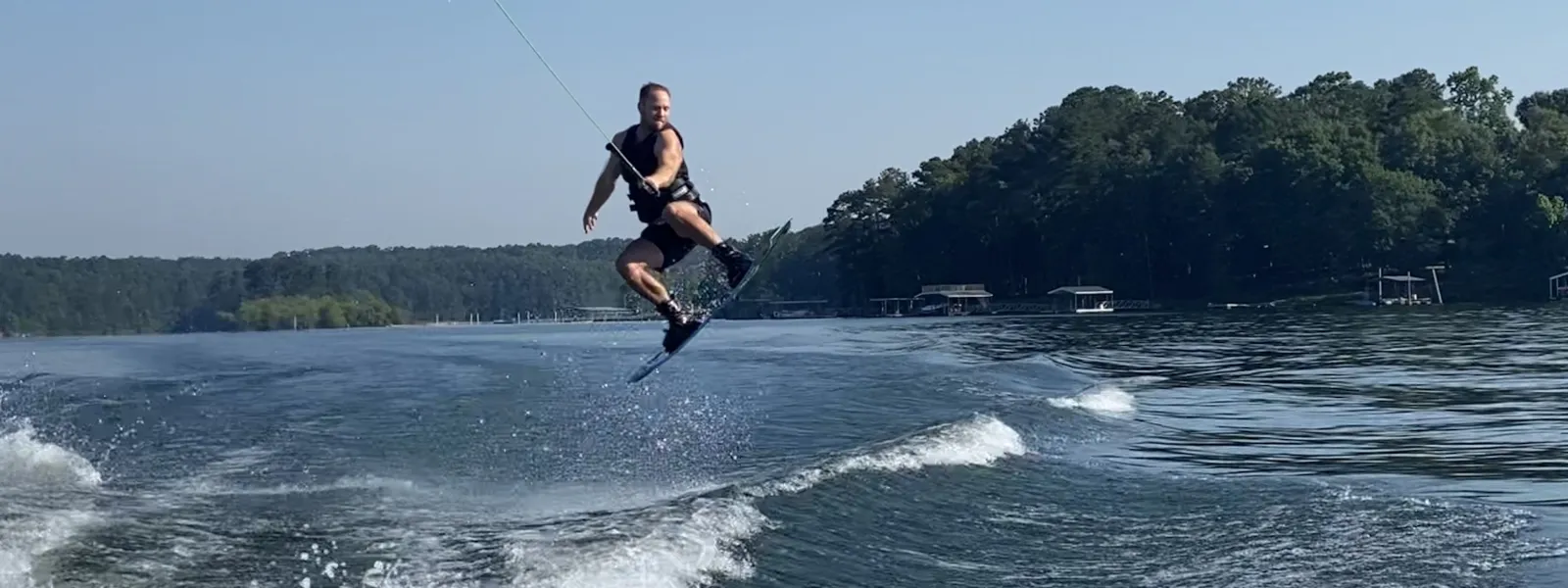 Man wakeboarding over lake water with forested shoreline under clear blue sky