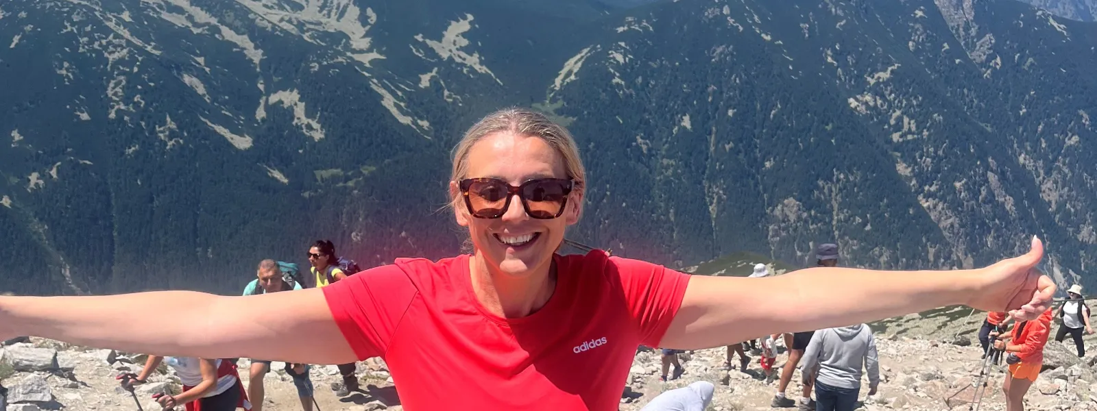 Smiling woman in red shirt with arms outstretched on rocky mountain trail with hikers and mountain range backdrop.