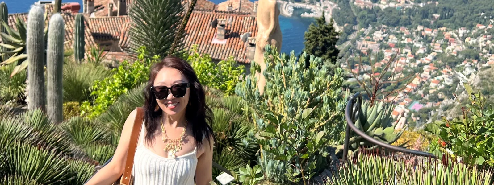 Woman in white dress and sunglasses standing in lush garden with French flag and coastal view in background