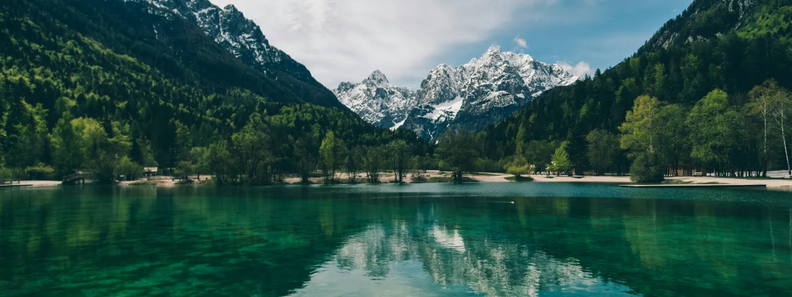 Clear turquoise lake with forested shores and snow-capped mountain peaks under a partly cloudy sky.