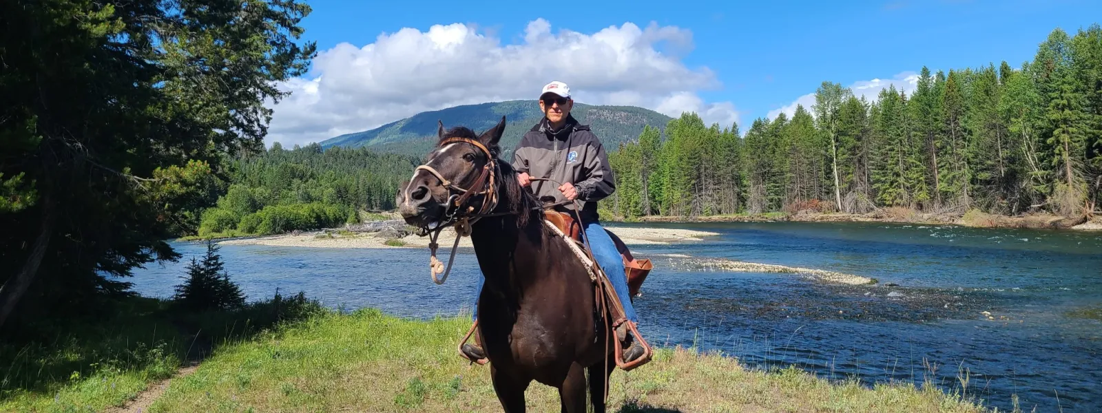 Person riding a black horse along a river with pine trees and mountains under a bright blue sky.