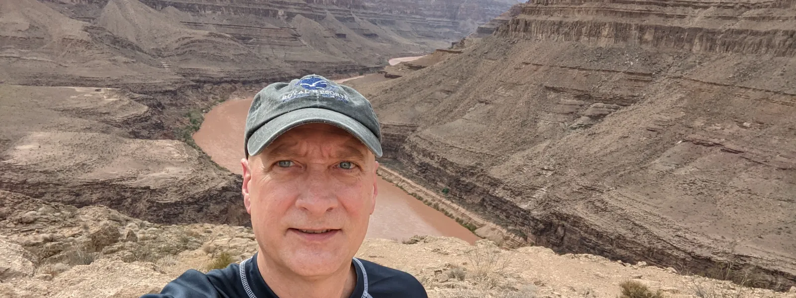 Man in black shirt and cap taking selfie at a scenic, rocky canyon with a winding river below under a cloudy sky.