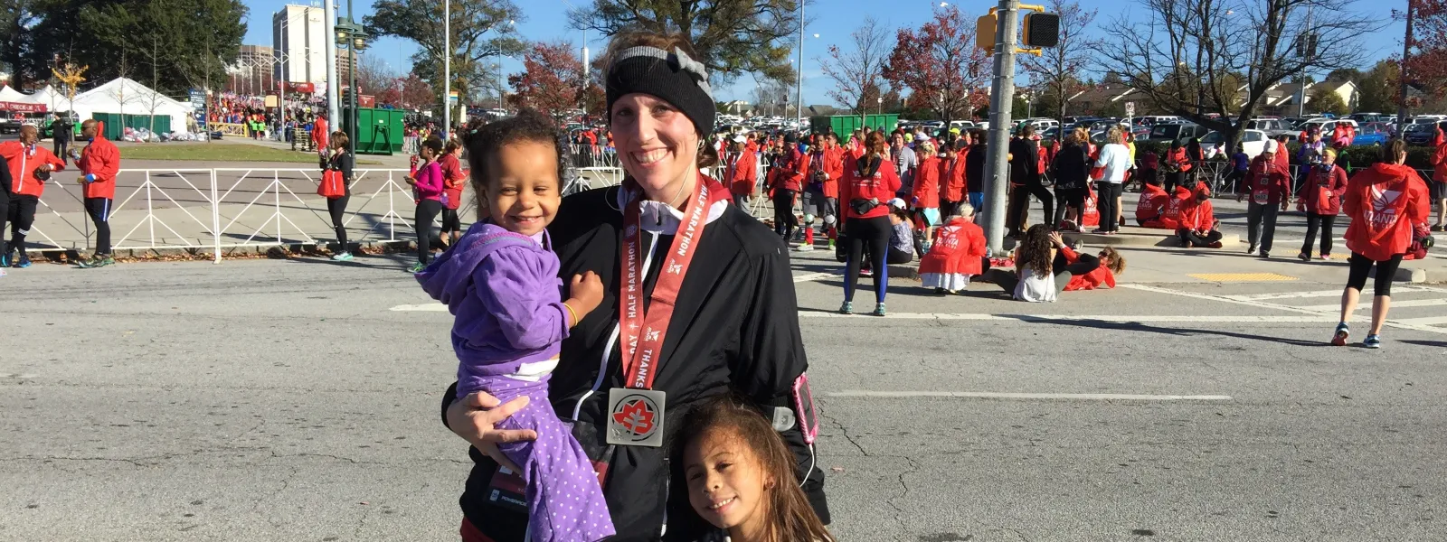 Smiling woman with two children at a race event wearing a finisher medal and winter clothing on a sunny day.