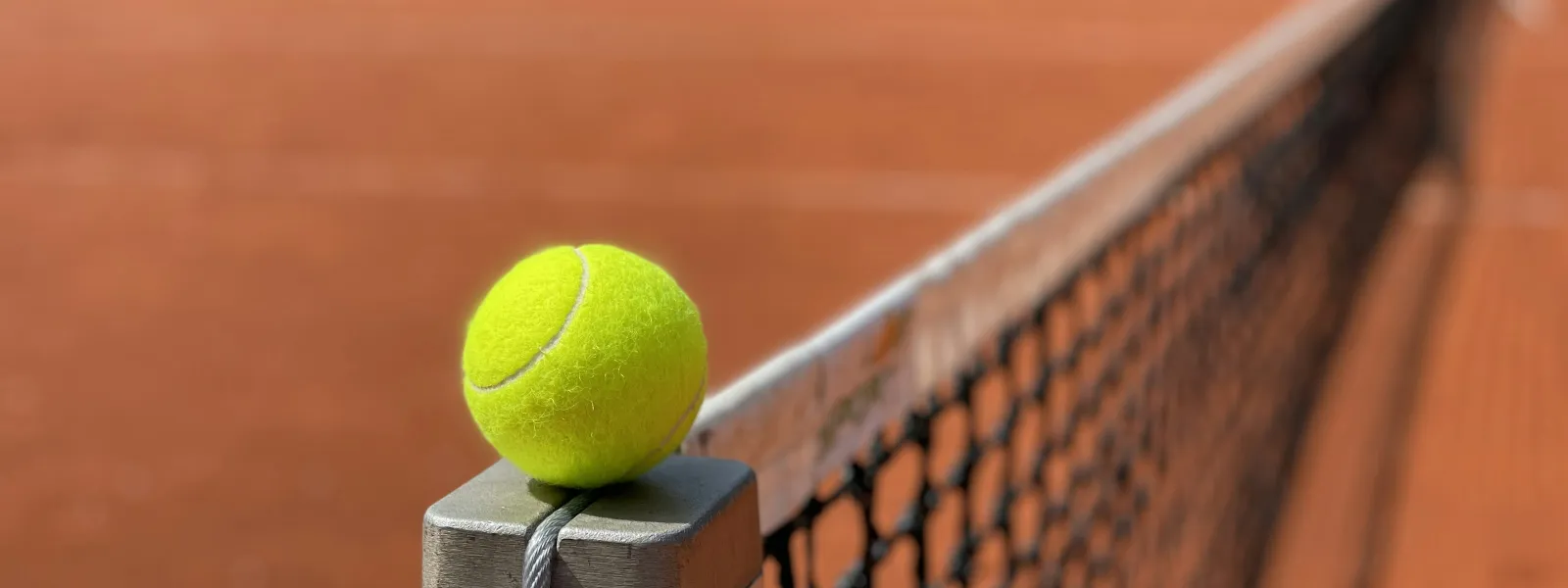 Bright yellow tennis ball resting on a metal net post at the center of a clay tennis court.
