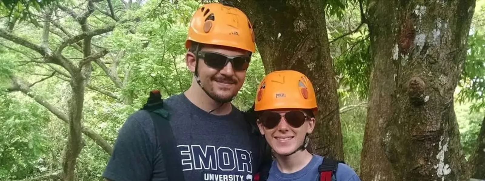 Couple wearing orange helmets and safety harnesses posing outdoors near large tree for ziplining or climbing.