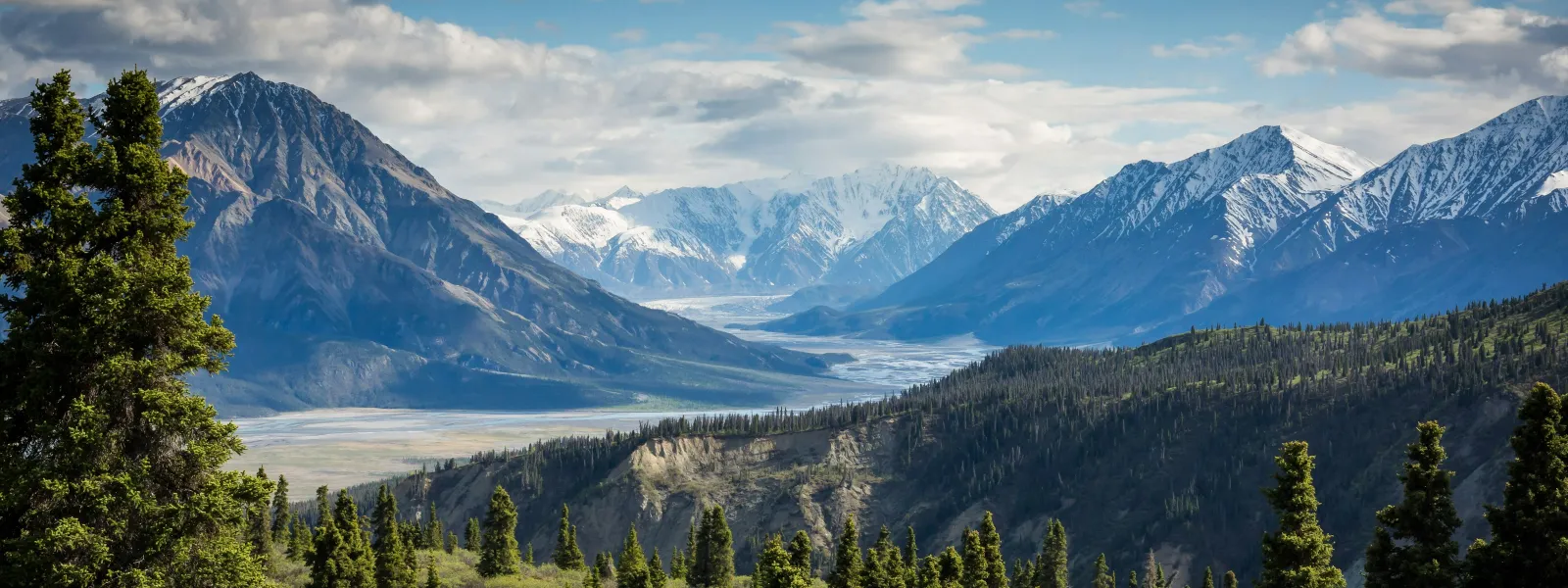 Panoramic view of snow-capped mountains with lush evergreen forest and partly cloudy sky in the background