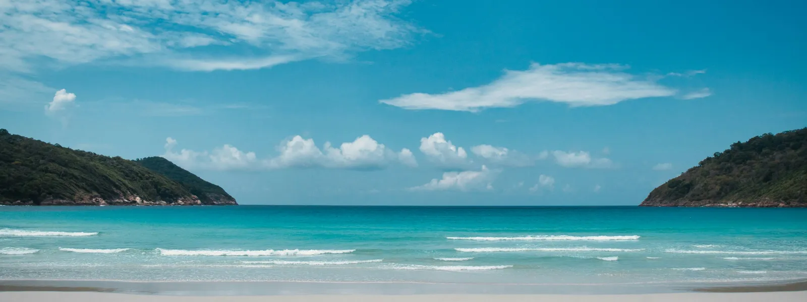 Serene beach with white sand, turquoise ocean waves, green hills, and partly cloudy blue sky.