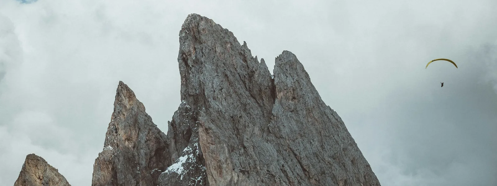 Jagged mountain peaks rise above grassy slopes under a cloudy sky with a paraglider flying nearby.