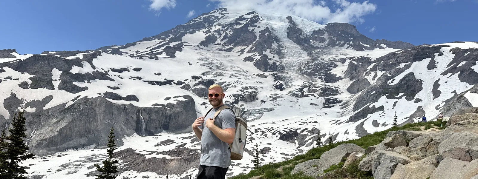 Hiker wearing shorts and backpack stands on rocky trail with snow-covered mountain and blue sky in the background
