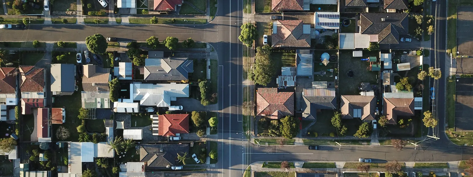 Aerial view of a suburban neighborhood with houses, streets, trees, and parked cars in daylight.