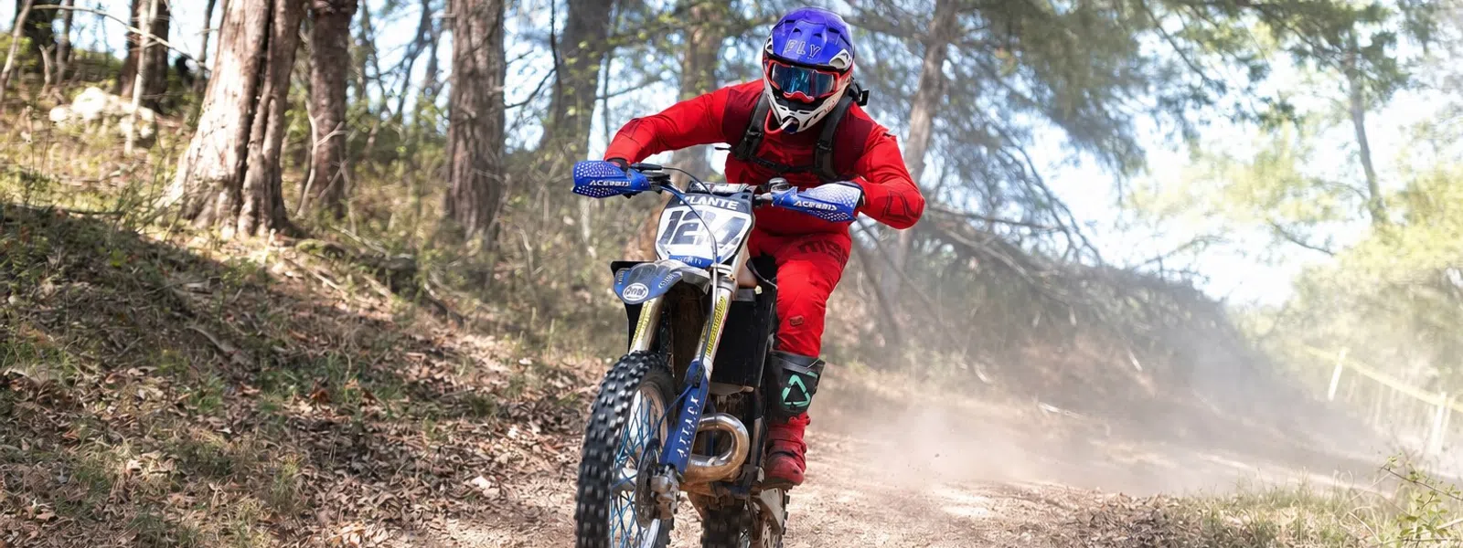 Dirt bike rider in red gear jumps over trail in forested off-road path during daytime.