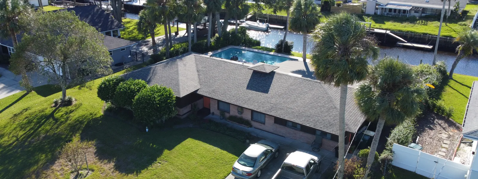 Aerial view of a residential house with a driveway, two parked vehicles, palm trees, and a backyard pool beside a canal.