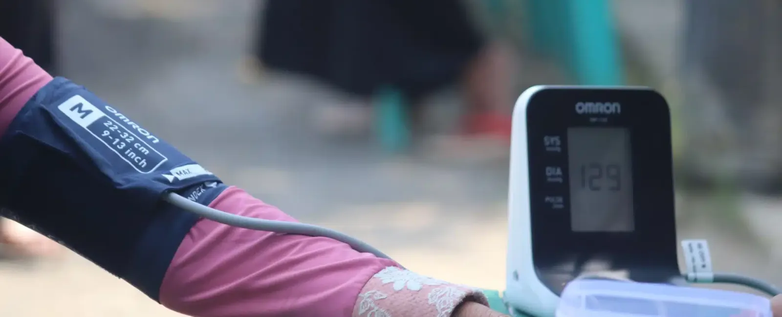 Person wearing a blood pressure cuff connected to an Omron monitor reading 129 mmHg sitting on a table outdoors.