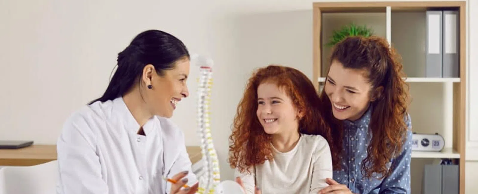 Pediatric doctor explaining spinal health to a smiling child and her mother in a clinic setting.