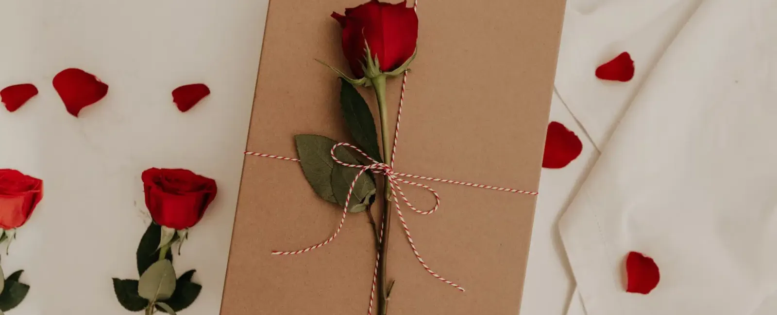 Brown gift box tied with string and topped with a single red rose on white fabric with scattered rose petals.