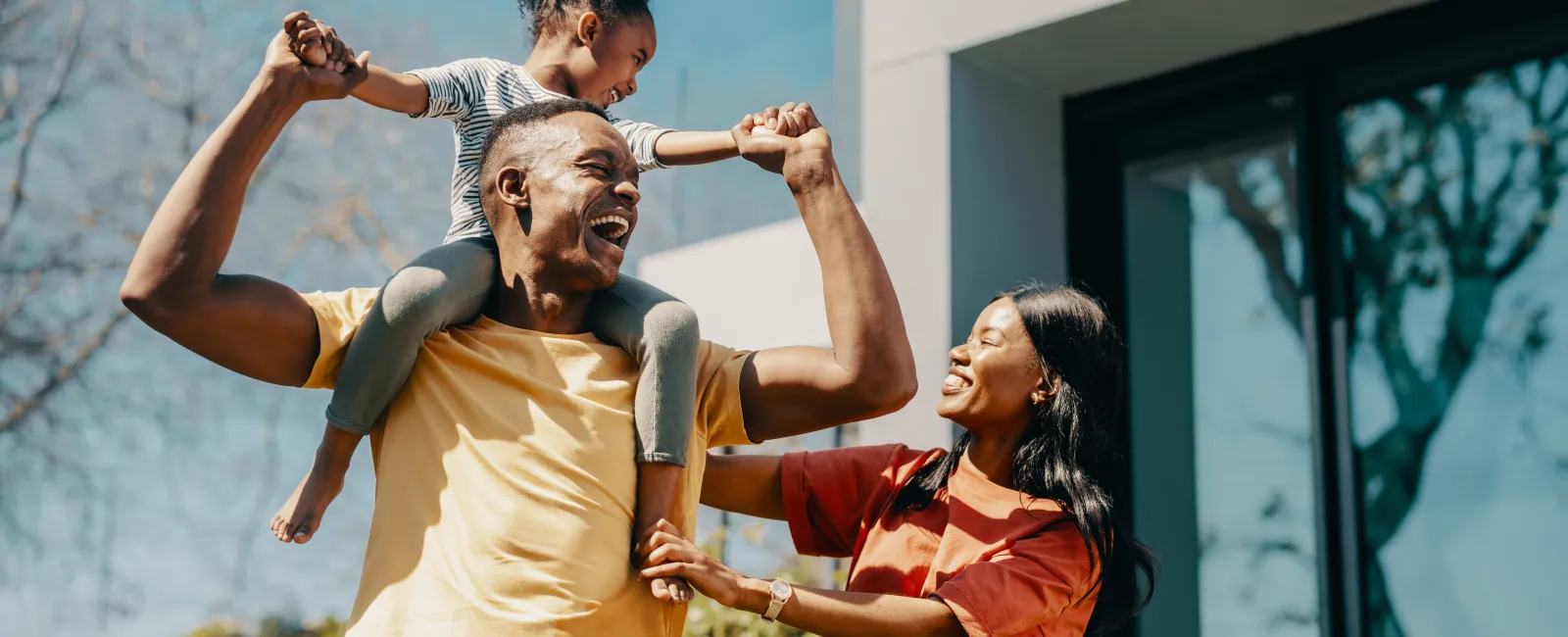 Happy family outdoors with father carrying daughter on shoulders and mother smiling nearby in sunny day.