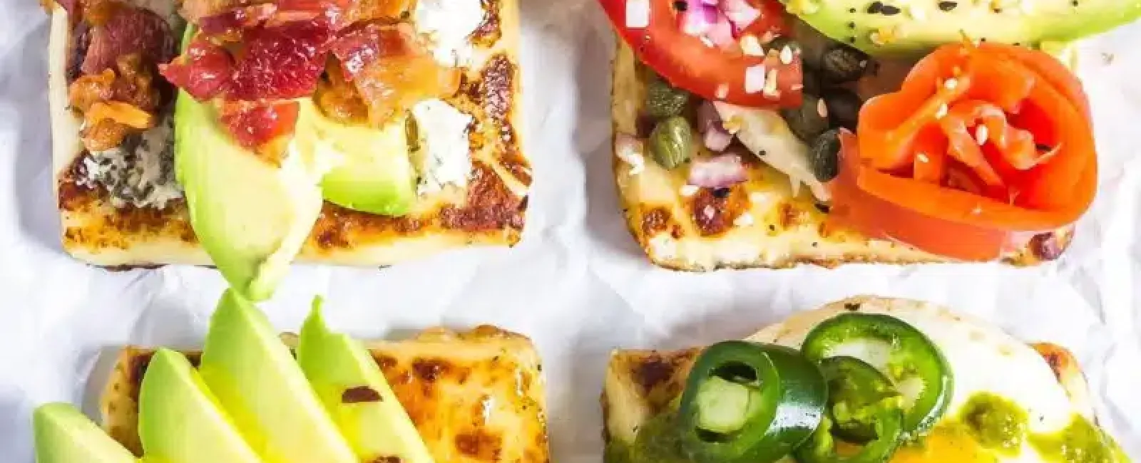 Four square toasts topped with avocado, bacon, tomato, egg, radish, and herbs on white parchment.
