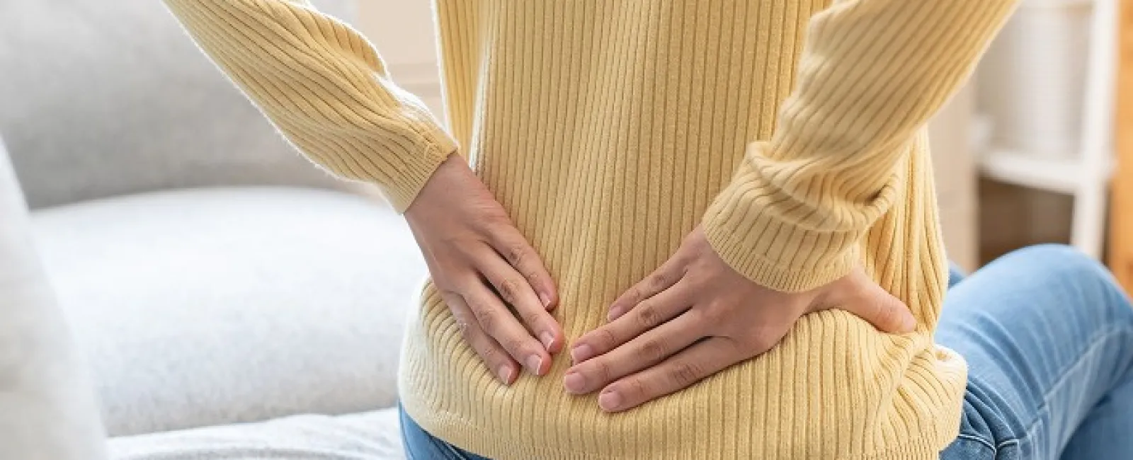 Person sitting on a couch holding their lower back, indicating back pain or discomfort indoors.