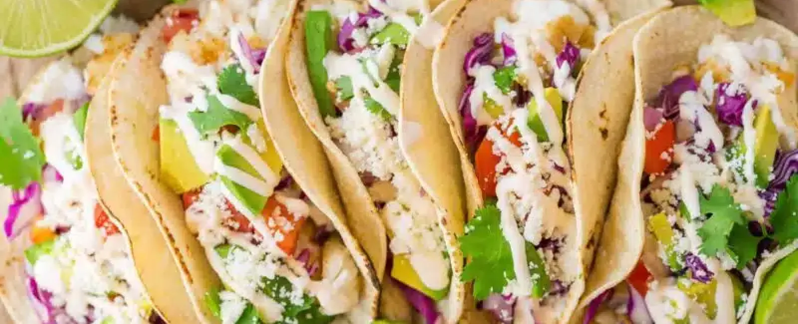Six fresh fish tacos with avocado, cilantro, purple cabbage, lime wedges, and creamy sauce on brown paper.