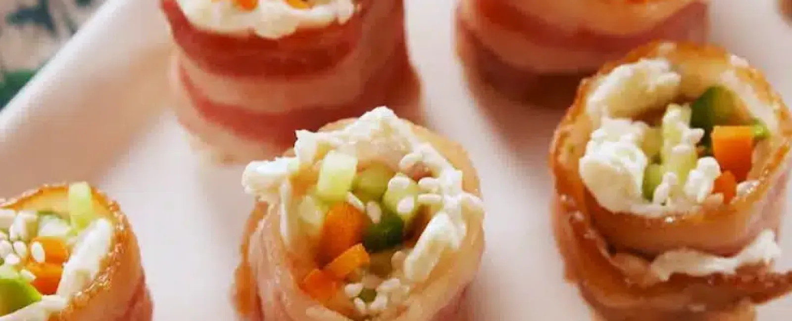 Bacon-wrapped sushi rolls with rice, cucumber, carrot, and cream cheese on white plate.