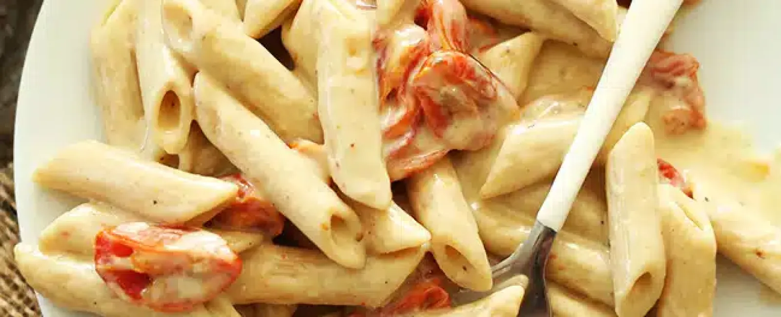 Creamy penne pasta with cherry tomatoes and herbs on a white plate with a fork.