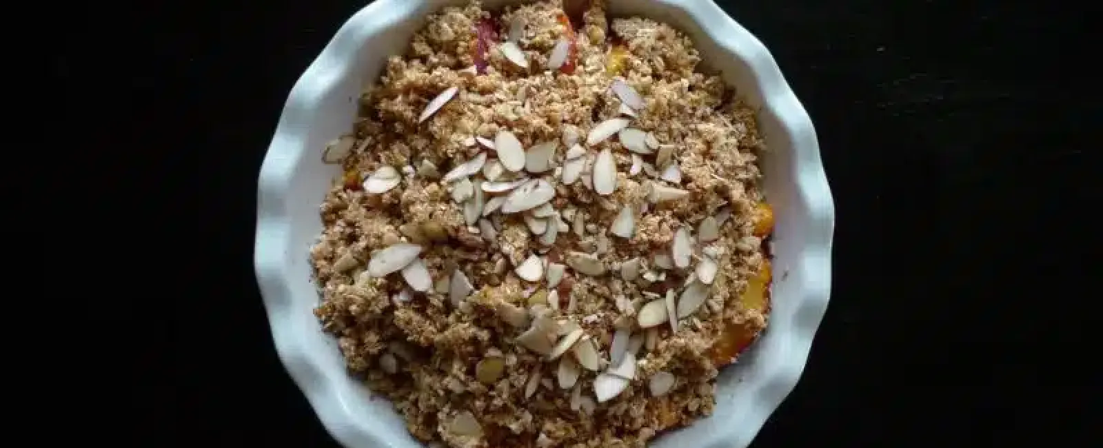 Top view of a fruit crumble dessert topped with sliced almonds in a white ceramic dish on black surface