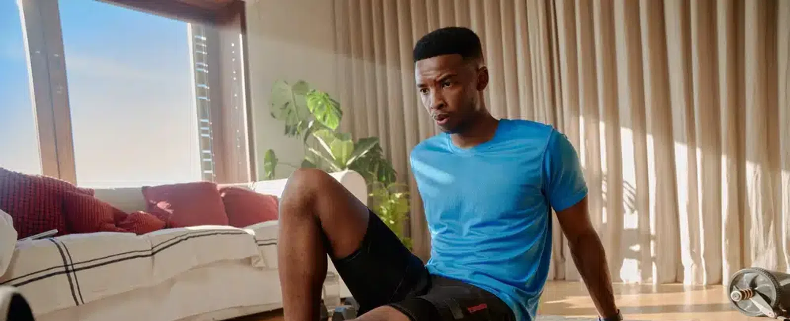 Man in blue shirt using foam roller on leg while exercising indoors on a yoga mat in bright living room