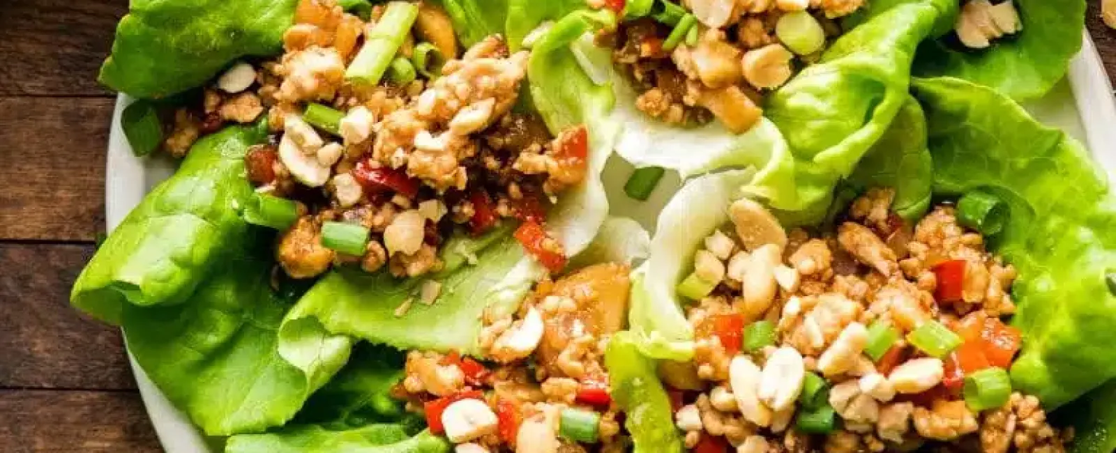 Fresh lettuce wraps filled with ground meat, chopped peanuts, red peppers, and green onions on a white plate.