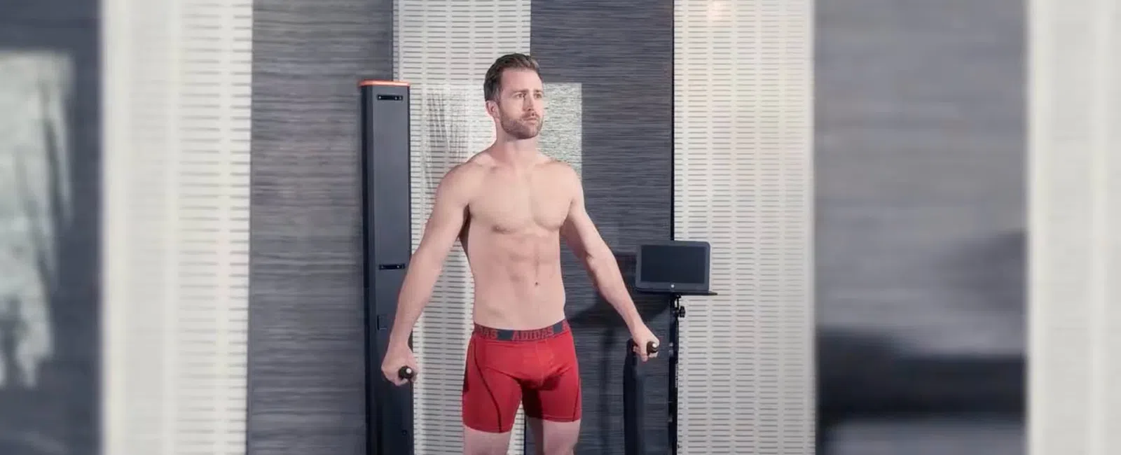 Man in red shorts using fitness equipment in a modern indoor gym with grey walls and digital screens.