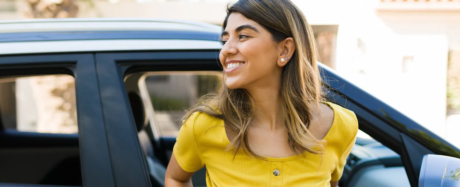 Smiling woman in yellow shirt leaning out of car window with urban background bright daylight.
