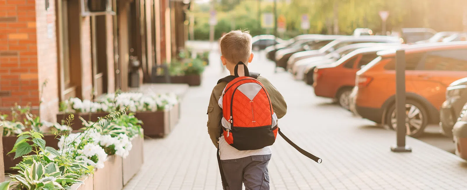 Young boy walking along city sidewalk with red backpack on sunny day near parked cars and planters.