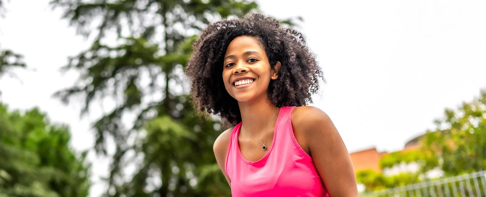 Smiling young woman in pink athletic wear outdoors with green trees and blurred background on a sunny day