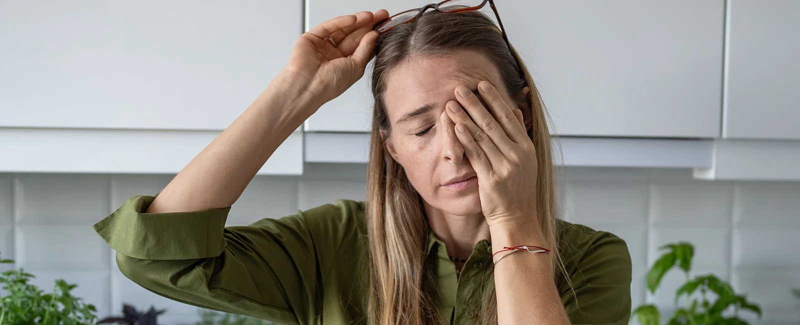 Woman with blonde hair in green shirt holding her forehead, appearing tired or stressed in a bright kitchen.