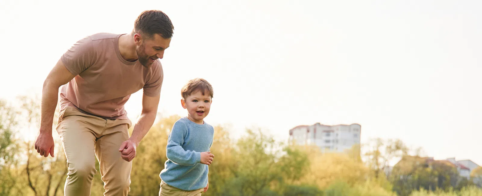 Father and young son running together in a sunny park with trees and distant buildings in the background