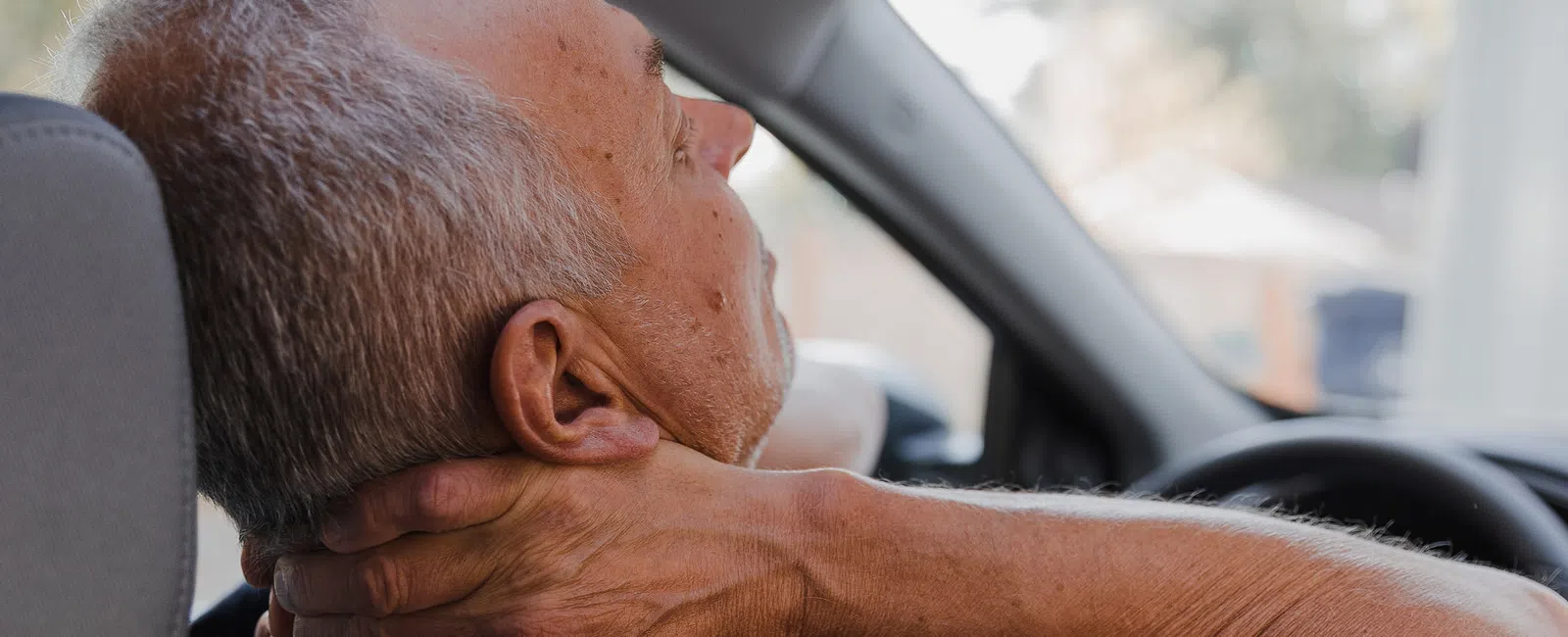 Senior man sitting in car holding neck with hand, showing discomfort or pain while driving.