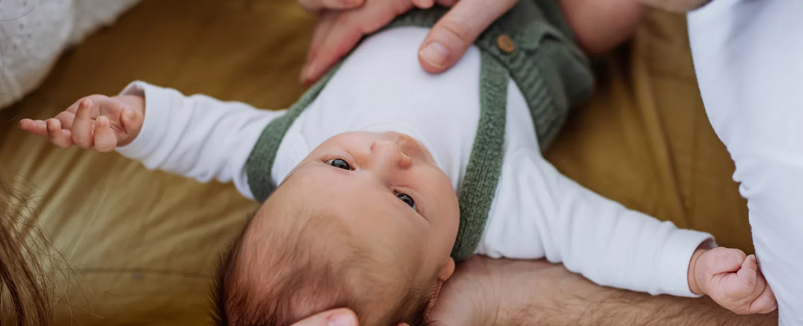 Baby lying on bed wearing white and green clothing, held gently by adults with blurred background.