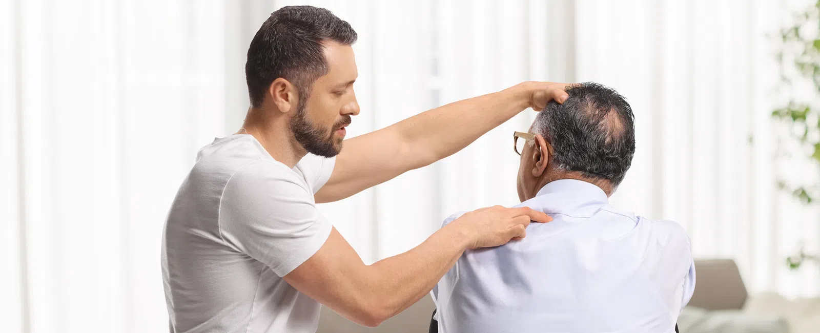 Young male chiropractor examining elderly man's neck during a therapy session in a bright home setting