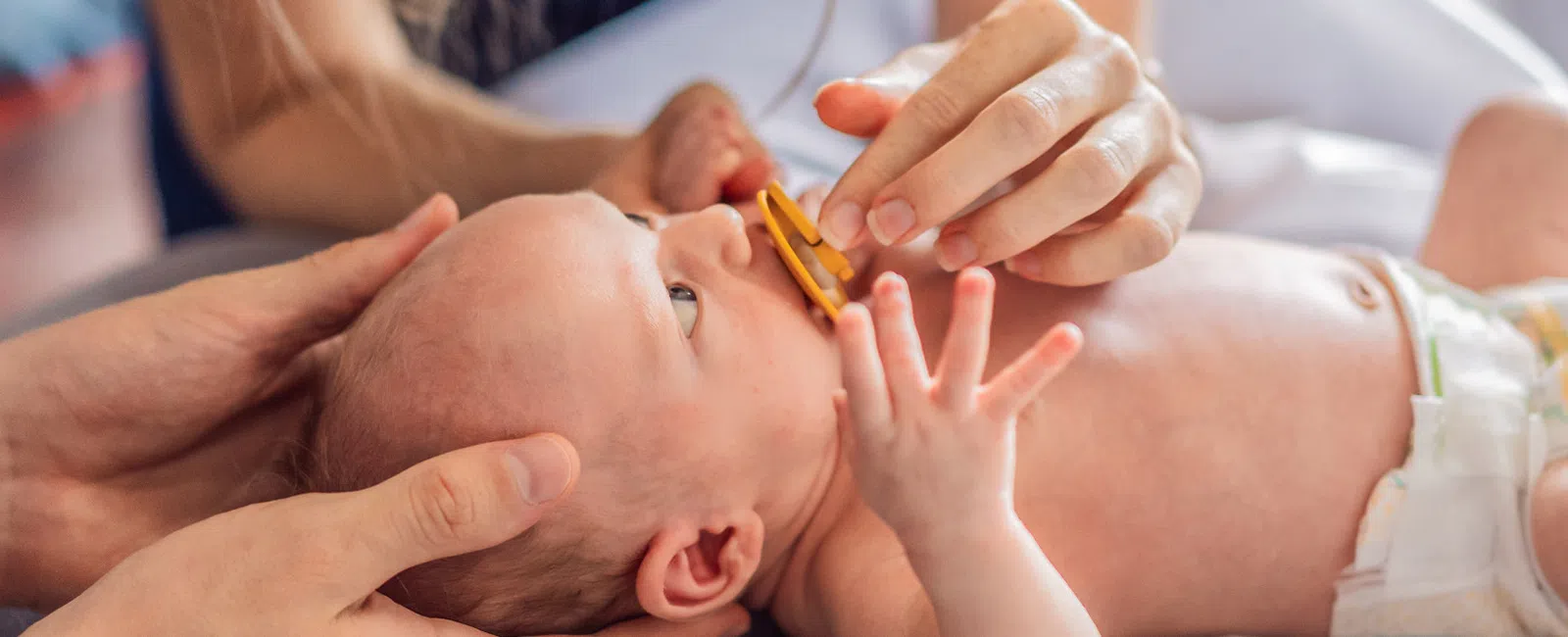 Newborn baby lying down while parents gently hold and feed with a pacifier at home.