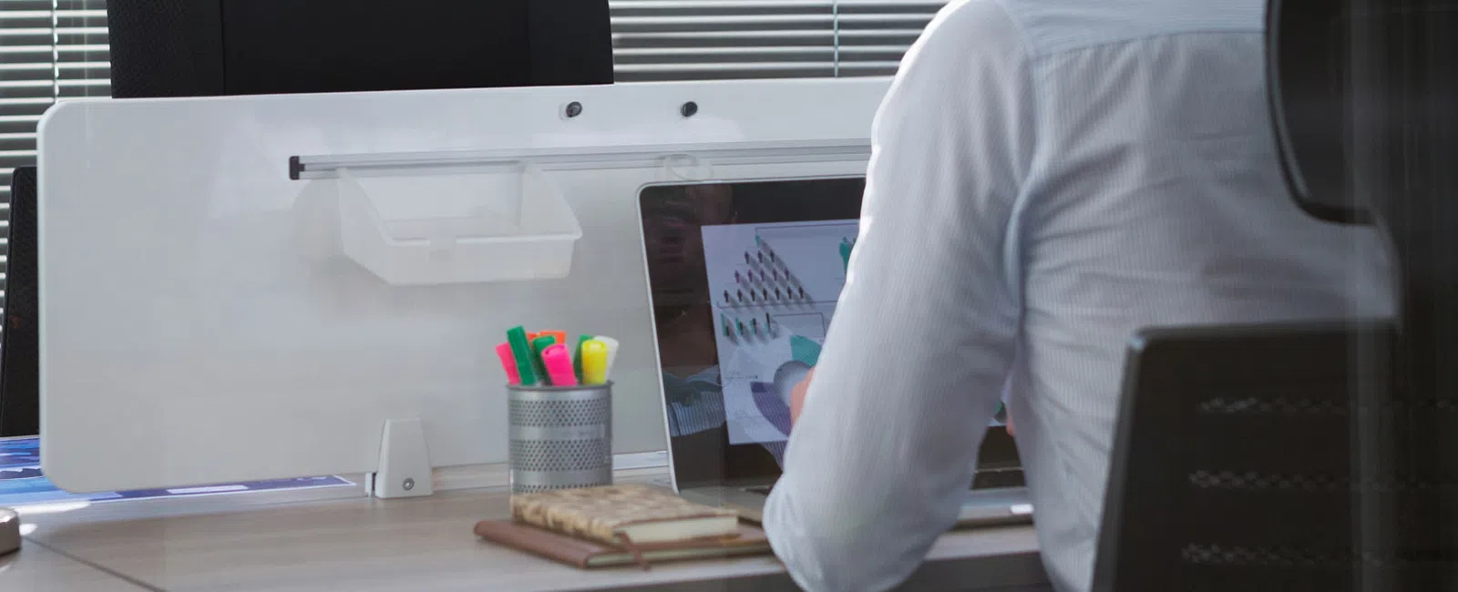 Person working on a laptop sitting in an ergonomic chair at a desk with office supplies, a notebook, and a modern lamp visible.