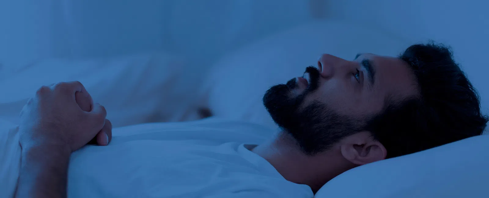 Man lying awake in bed with hands clasped, looking up, surrounded by a blue-toned dimly lit room.
