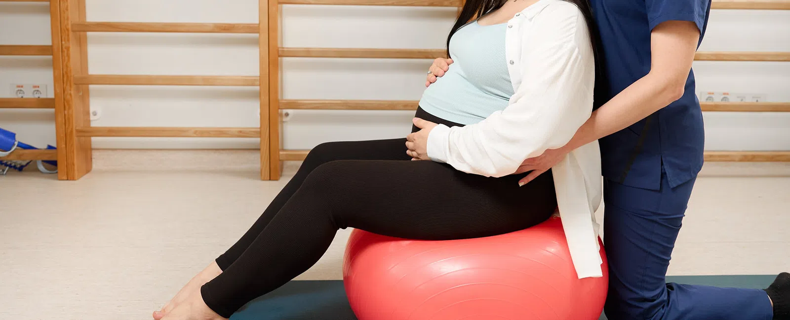 Pregnant woman sitting on exercise ball with therapist support in a physical therapy room.