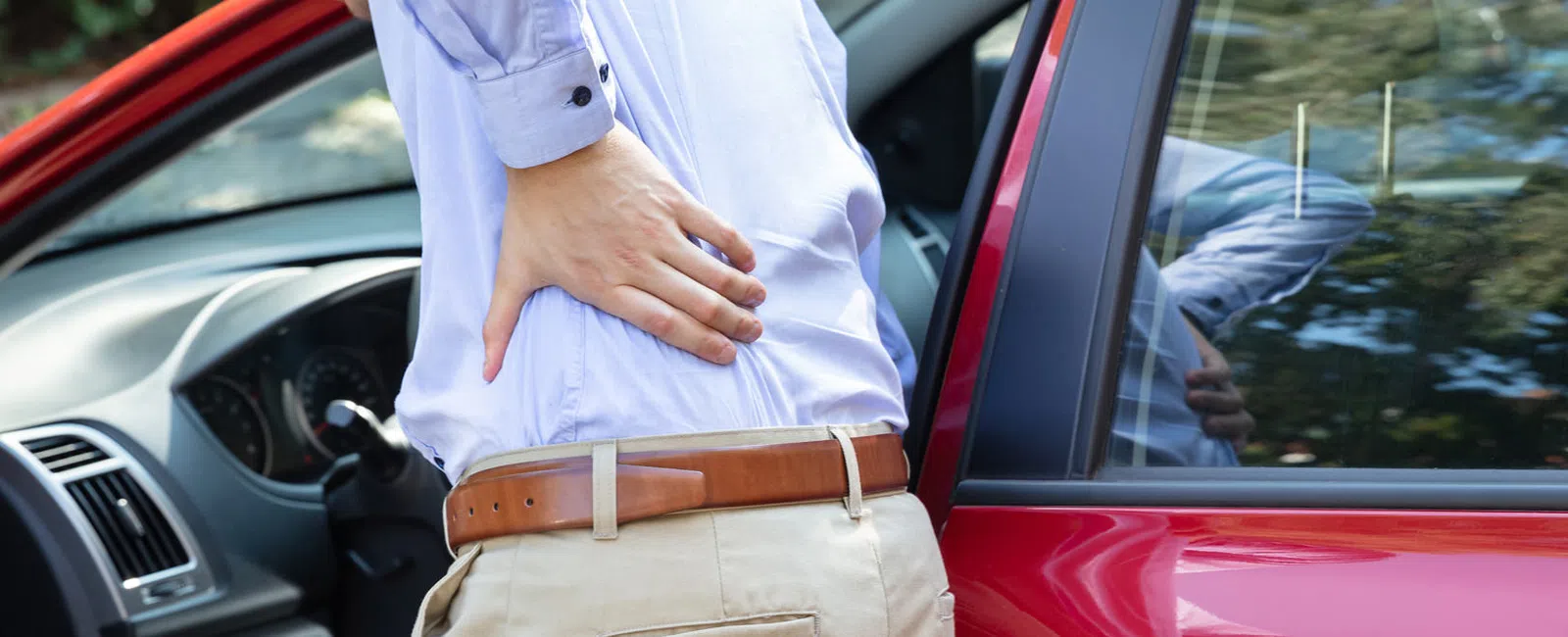 Person holding lower back in pain next to open red car door, indicating discomfort after driving.