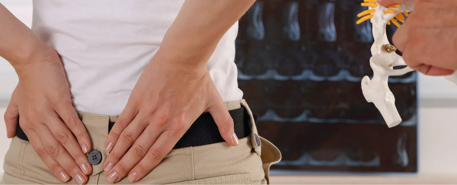 Person holding lower back in pain as chiropractor shows spine model with X-ray in background.