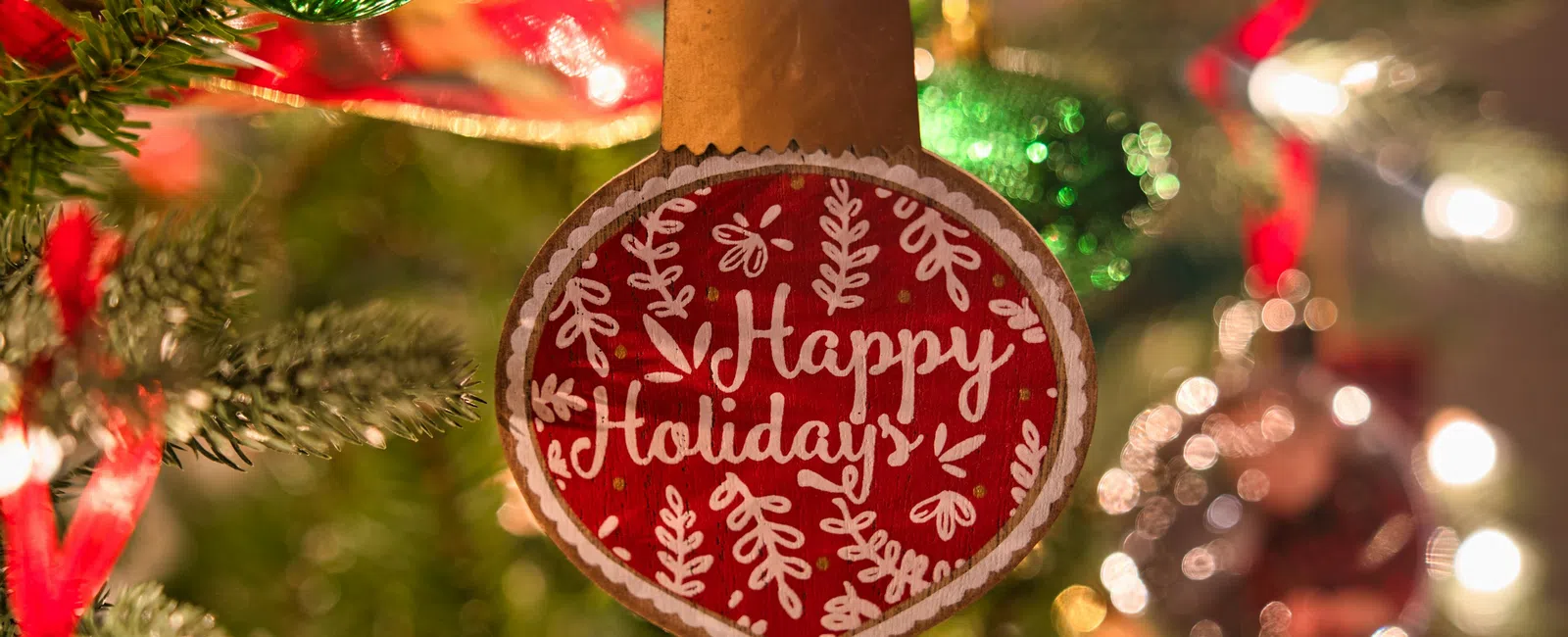 Close-up of a red holiday ornament with Happy Holidays text hanging on a decorated Christmas tree with lights.