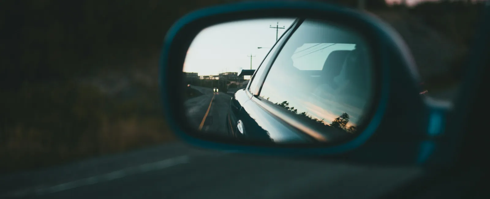 Car side mirror reflecting a road and telephone poles during sunset with approaching headlights.