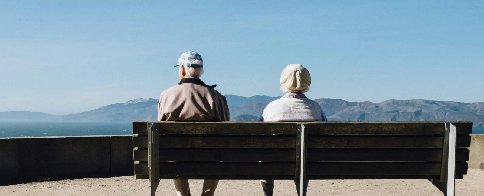 Two elderly people sitting on a bench facing mountains under a clear blue sky on a sunny day