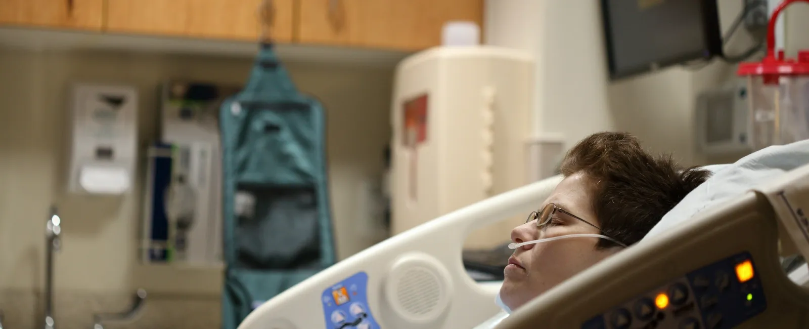 Patient lying in hospital bed receiving oxygen with medical equipment and cabinets in background.