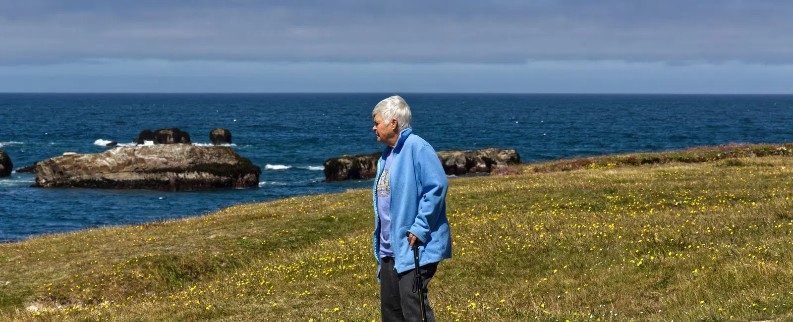 Elderly woman with cane standing on grassy seaside cliff under clear sky near ocean rocks.
