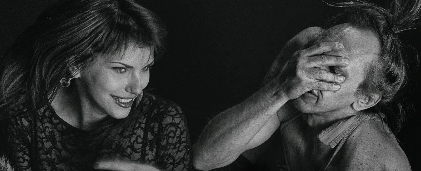 Black and white photo of smiling woman and man covering his face with his hand in dramatic lighting.